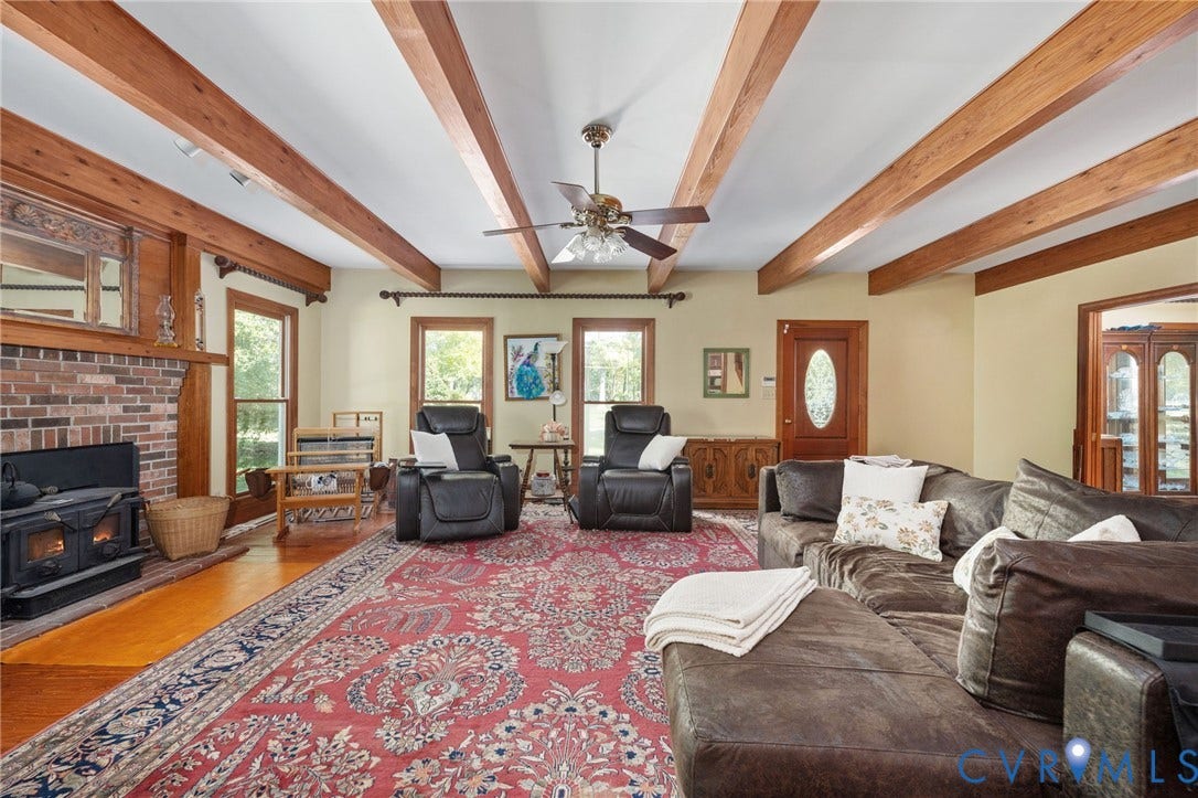 Exposed wood beam ceiling, brick fireplace with wood-burning stove, wood-finish flooring, multiple windows, and a ceiling fan