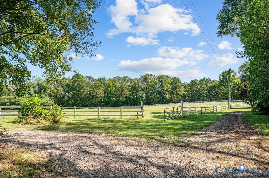 Expansive property featuring a gravel driveway, lush green lawn, white perimeter fencing, and mature trees