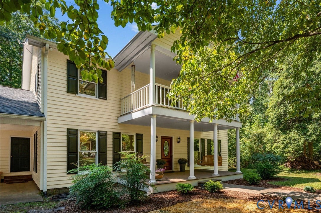 Two-story residence featuring siding exterior, an upper-level balcony, a covered front porch with columns, a dark wood-finish front door with an oval glass insert, and mature perimeter trees