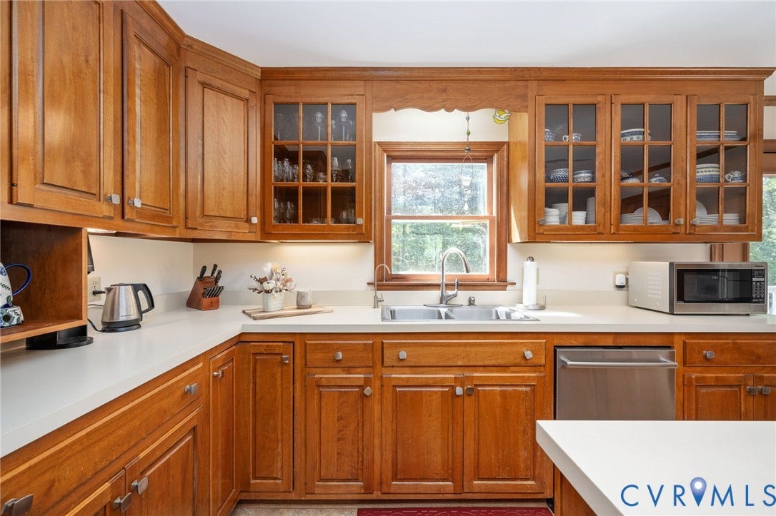 Wood-finish cabinetry with glass-front display cabinets, light-toned countertops, and a double basin sink with a gooseneck faucet