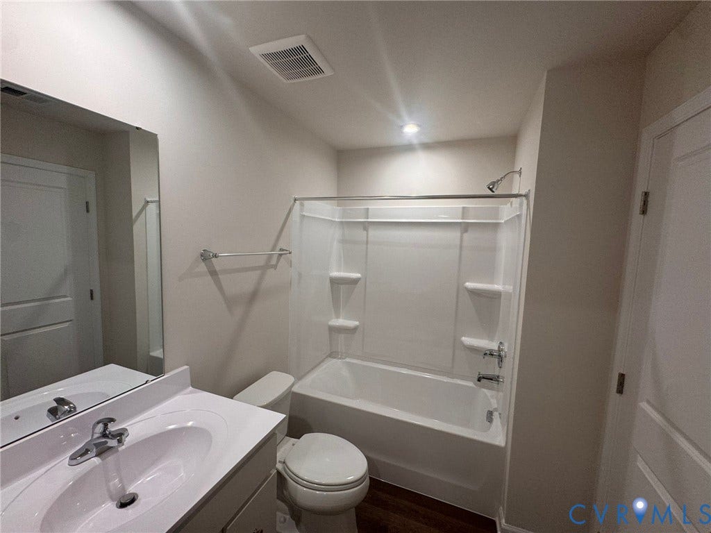 Bathroom featuring a vanity with an integrated sink, a large wall mirror, a toilet, a bathtub with an integrated shower, and wood-finish flooring