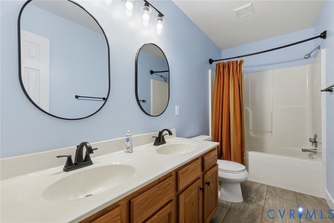 4350 Steger Creek Drive, Powhatan, VA 23139 Dual oval mirrors, double vanity with integrated sinks, wood-finish cabinetry, matte black fixtures, and a shower-tub combination