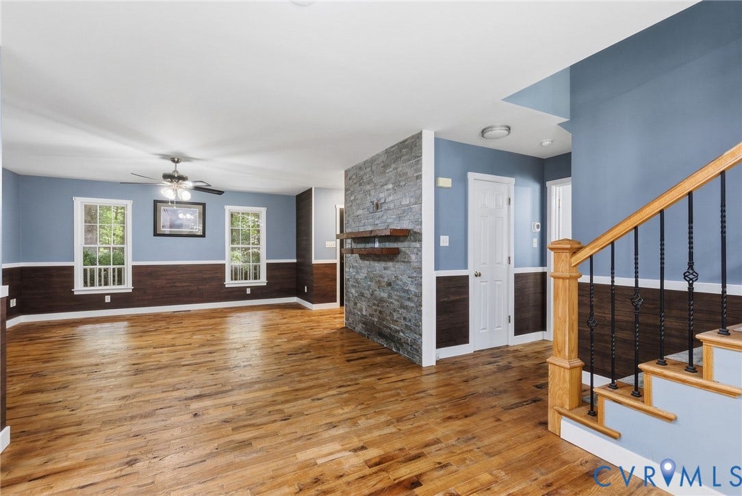 4350 Steger Creek Drive, Powhatan, VA 23139 Wood-finish flooring throughout, stacked stone accent wall with floating shelves, wood-paneled wainscoting, classic spindle staircase with wood handrail, and a ceiling fan with light fixture