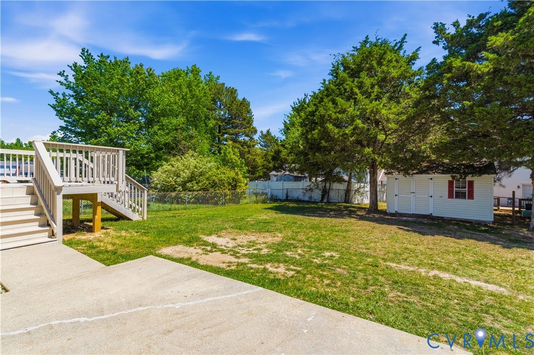 952 Pleasant Street, Henrico, VA 23223 Expansive backyard featuring a concrete patio, wood deck with stairs, chain-link fencing, mature trees, and a white storage shed with red shutters