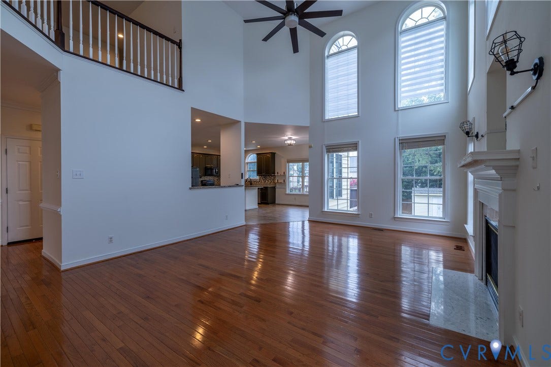 Soaring ceilings with transom windows, wood-finish flooring, a decorative fireplace with a stone hearth, and an upper-level balcony with white balusters and a dark wood handrail