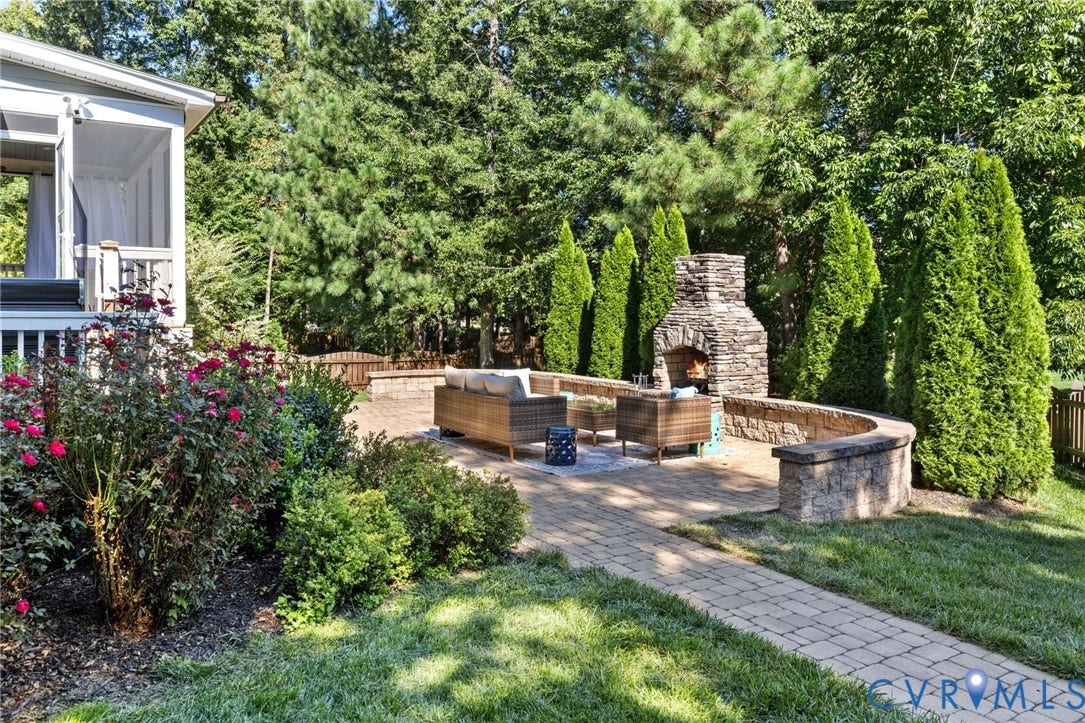 Expansive paver patio featuring a stone fireplace, curved stone retaining wall, and verdant landscaping