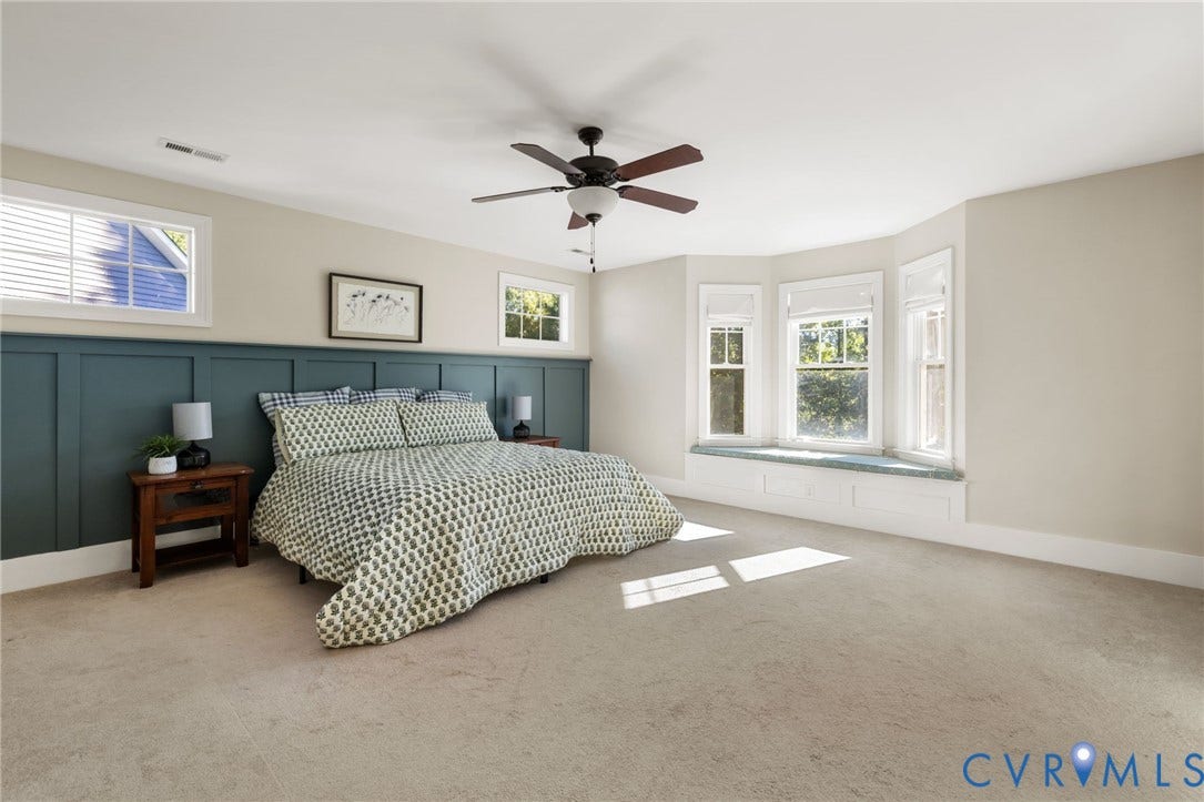 Expansive room featuring a bay window with integrated bench seating, wainscoting accent wall, ceiling fan, and light-toned carpeting