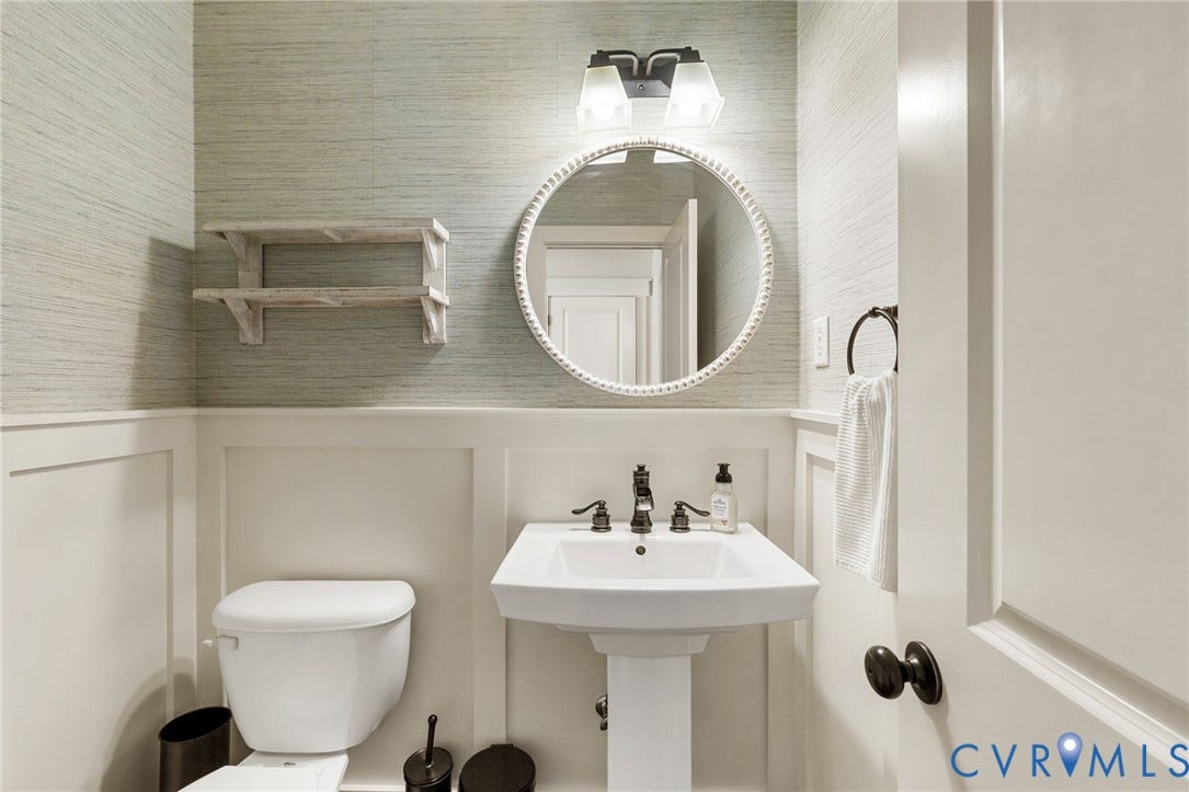 Pedestal sink with dark-finished faucet, wall-mounted vanity lighting, round mirror, and wainscoting with textured wall covering