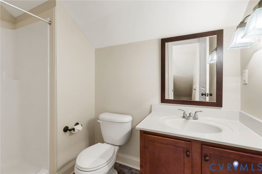 White shower surround, white toilet, wood-finish vanity with white countertop and integrated sink, framed mirror, and wall-mounted light fixture