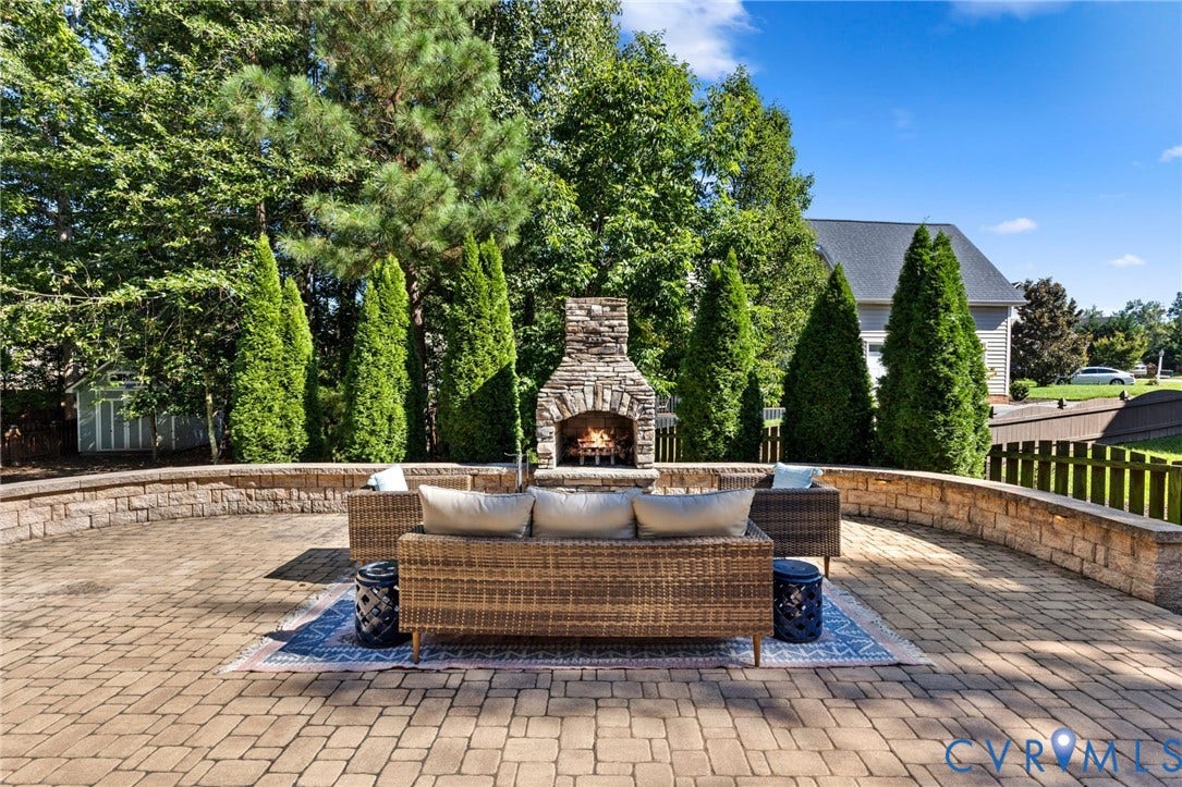 Expansive paver patio with a built-in stone fireplace, surrounded by a stone retaining wall and mature evergreen landscaping