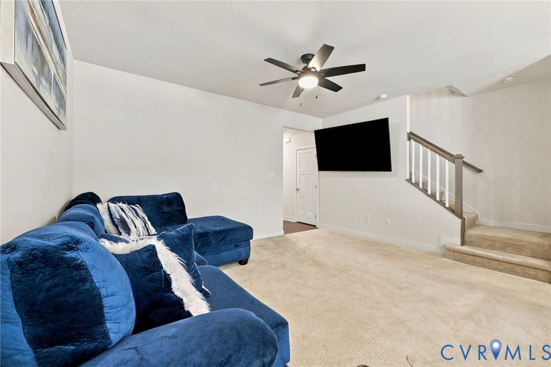 Neutral carpet flooring, white walls, ceiling fan with integrated lighting, wall-mounted television, and a staircase with white balusters and wood-finish handrail