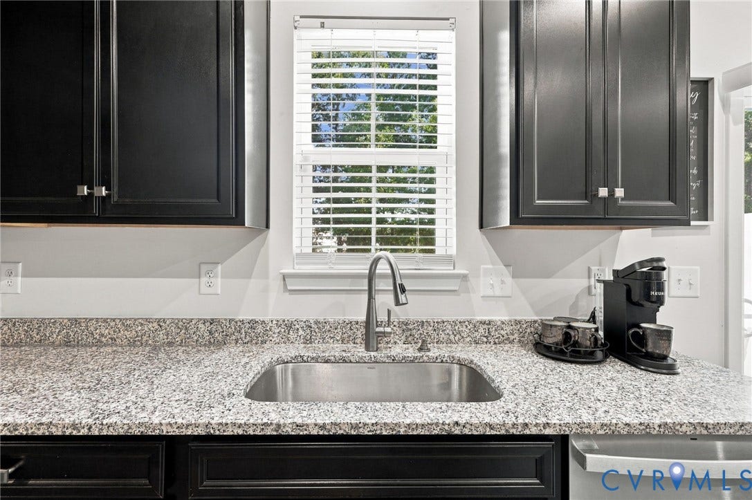 Stainless steel sink with a gooseneck faucet, light granite countertops, black cabinetry with square pull hardware, and a window with blinds