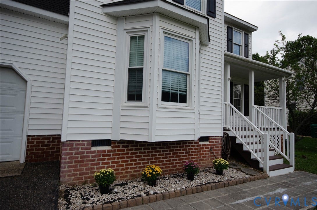White siding exterior with a prominent bay window, brick foundation, covered front porch, and a paver walkway