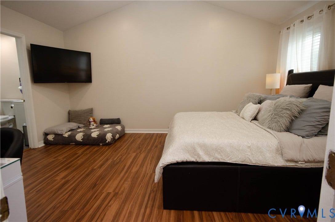 Vaulted ceiling, wood-finish flooring, wall-mounted television, window with white blinds, and a built-in light fixture