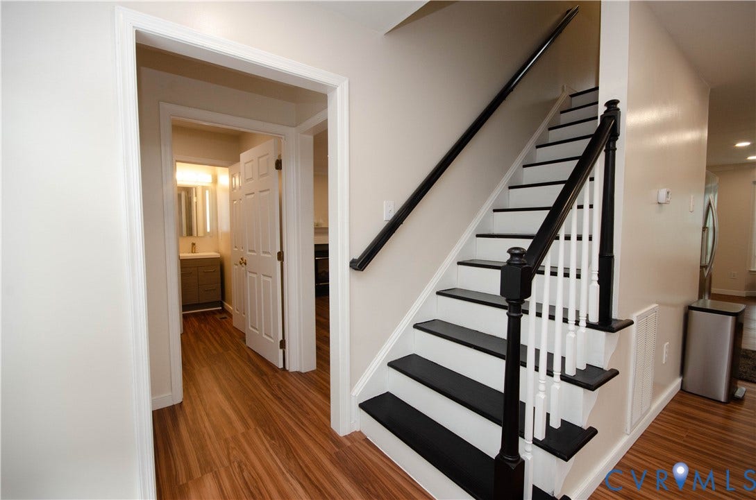 Wood-finish flooring throughout, two-tone staircase with dark treads and white risers, dark wood handrail, paneled interior door, and a bathroom vanity with a mirror and wall-mounted lighting
