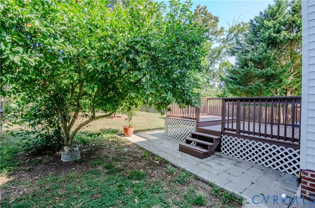 Elevated wood deck with railing and lattice skirt, paver patio, mature tree, and surrounding greenery
