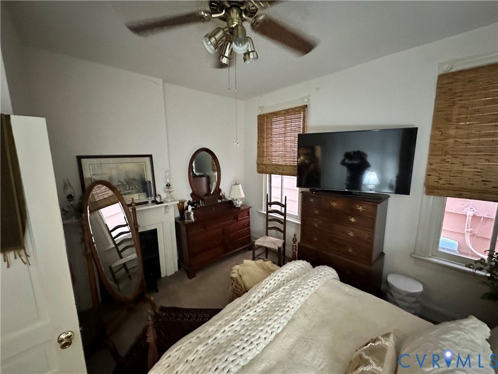 Ceiling fan with light fixture, white walls, two windows with woven shades, and a decorative fireplace surround