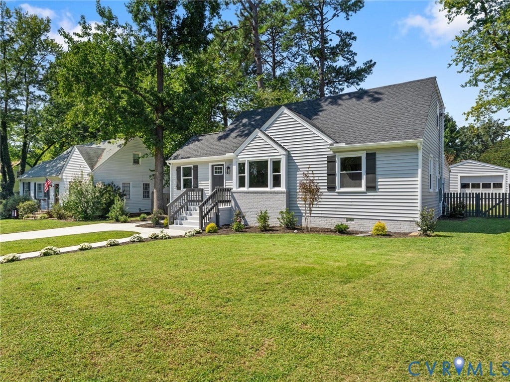 Light gray siding exterior with dark shutters, front porch with dark railings and steps, attached garage with a fence, mature trees, and a concrete driveway