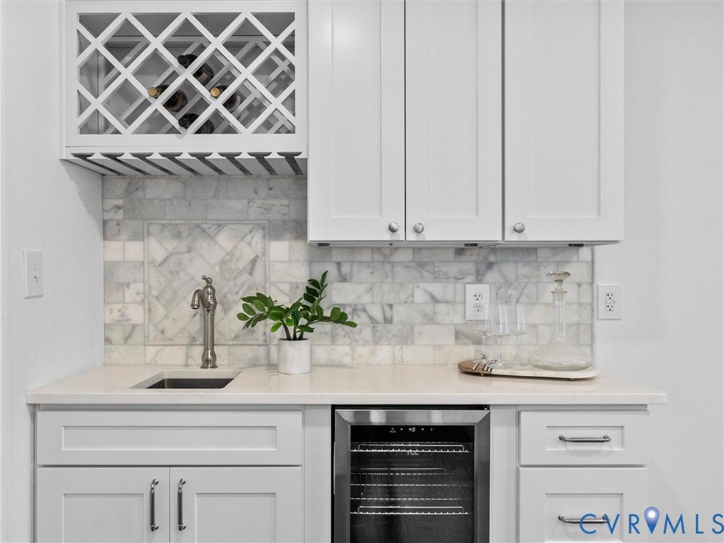 White cabinetry with contrasting hardware, marble-look tile backsplash, built-in wine rack with glass storage, integrated beverage cooler, and an undermount sink with a gooseneck faucet
