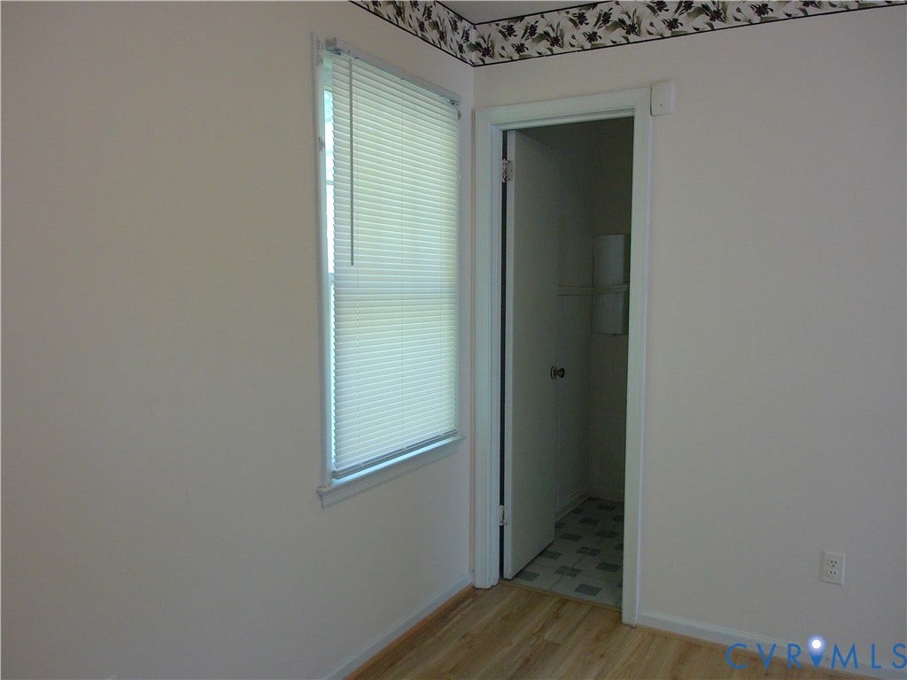 Wood-finish flooring with light-colored walls, a window with blinds, and a doorway featuring a dark door handle