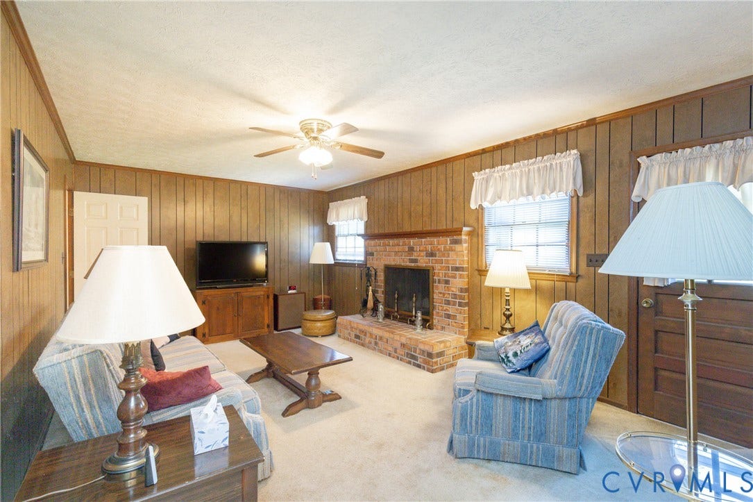 3640 Kiefer Road, Chester, VA 23831 Wood panel walls and wood-finish trim throughout, brick fireplace with hearth, ceiling fan with integrated lighting, and two windows with white blinds