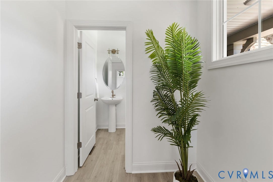 12506 Ashton Dell Road, Chester, VA 23831 White interior walls with a window providing natural light, wood-finish flooring, and a white pedestal sink with an oval mirror and wall-mounted light fixture