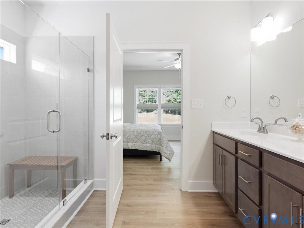 3102 Kensington Lane, Goochland, VA 23063 Bathroom featuring a dual vanity with white countertops, wood-finish flooring, a glass-enclosed shower with a built-in bench, and a transom window