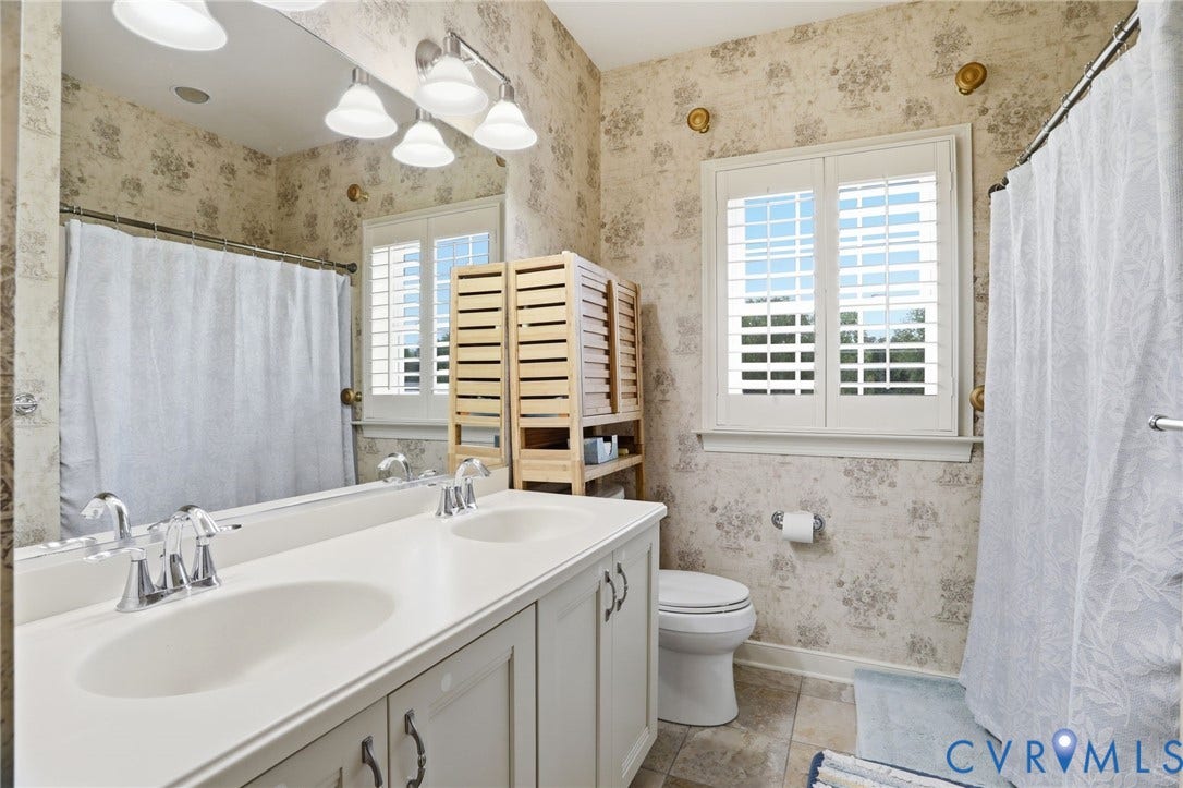 Dual vanity with integrated sinks and chrome fixtures, framed mirror, wall-mounted light fixture, louvered window shutters, and tiled flooring