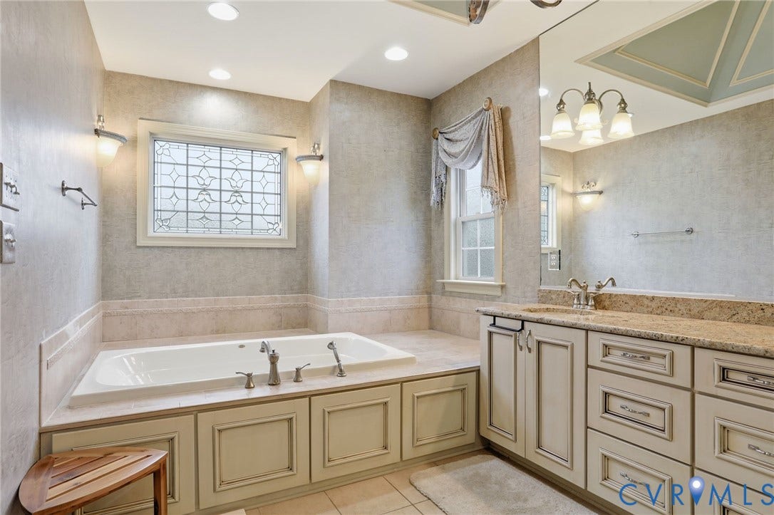 Built-in jetted tub with custom surround, leaded glass window, dual vanity with undermount sinks, and stone countertop