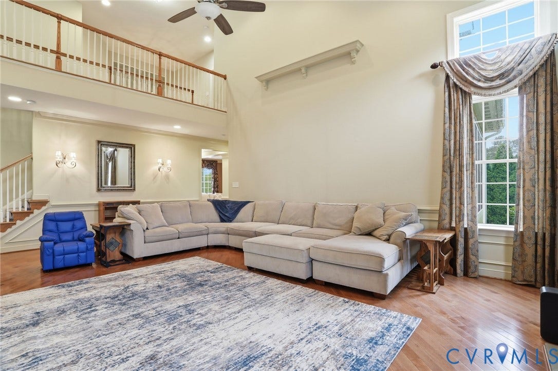 Two-story living area with hardwood flooring, ceiling fan, and large windows with custom window treatments