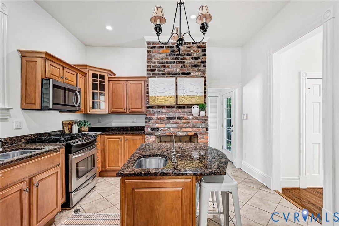 615 N 23rd Street, Church Hill Central, Richmond, VA 23223 Kitchen featuring wood cabinetry, stainless steel appliances, dark stone countertops, a central island with a sink, and a brick accent wall with an integrated fireplace