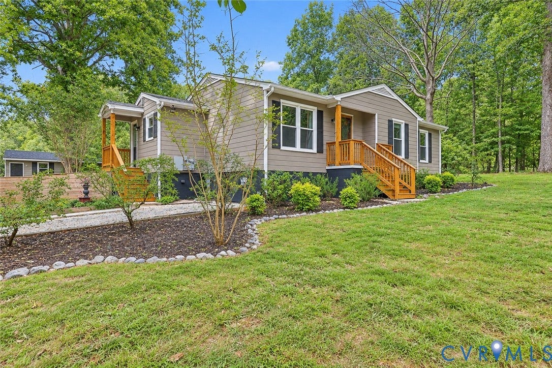 Neutral-toned siding exterior with dark shutters, natural wood-finish porch and railings, and a landscaped front yard with mature plantings
