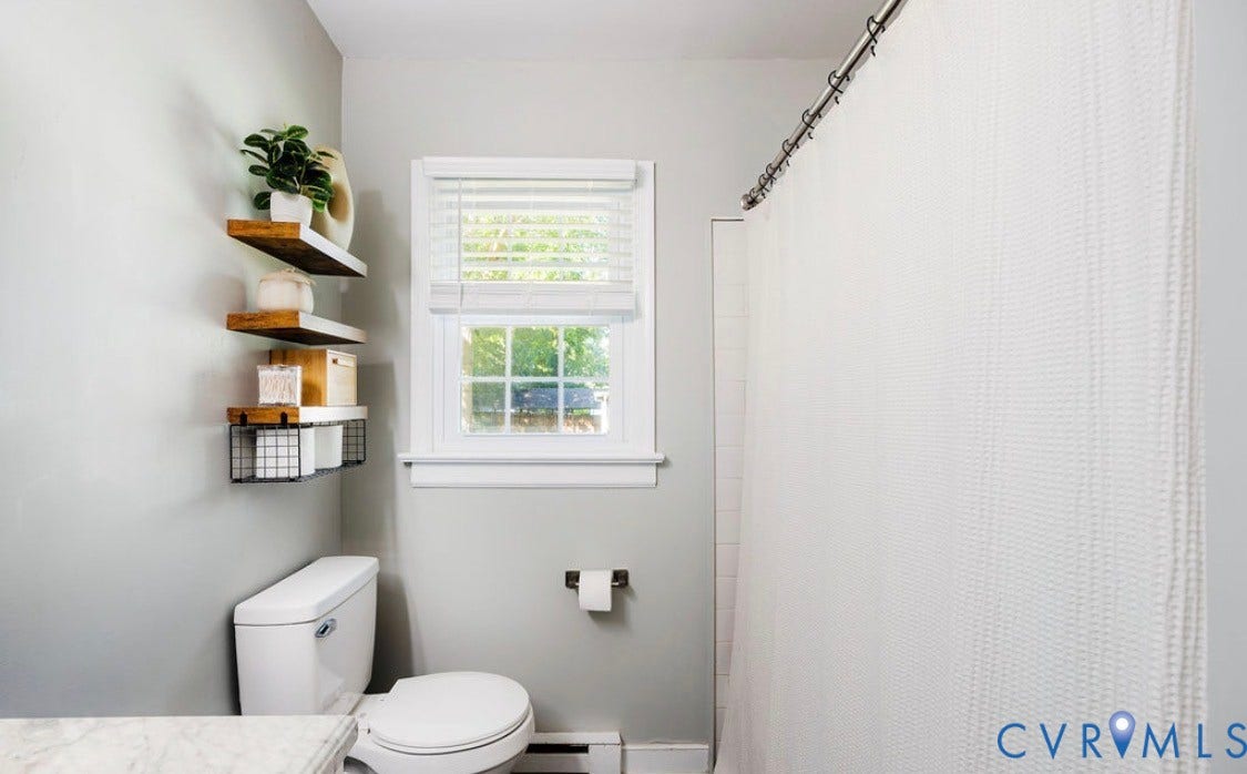 White ceramic toilet, window with blinds, wall-mounted shelving with wood-finish tops, shower area with white tile surround, and a curved shower rod