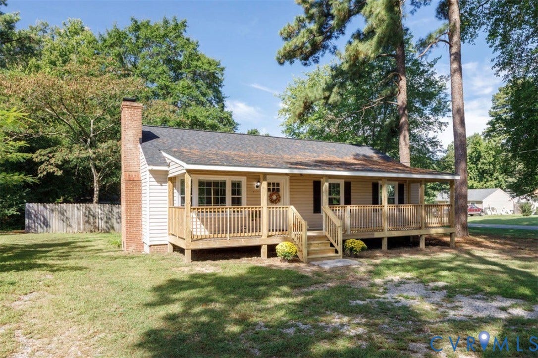Front porch with wood railing and steps, tan siding, dark shingle roof, and a brick chimney