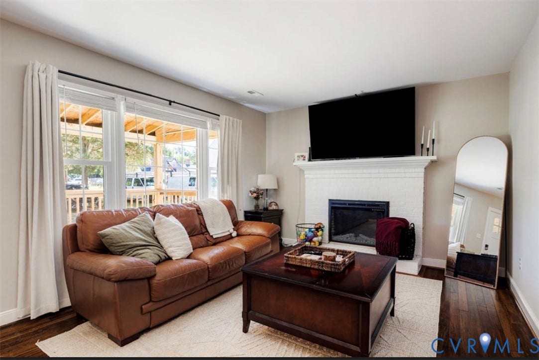 Living area with wood-finish flooring, a white brick fireplace with a mantel, and large windows providing natural light