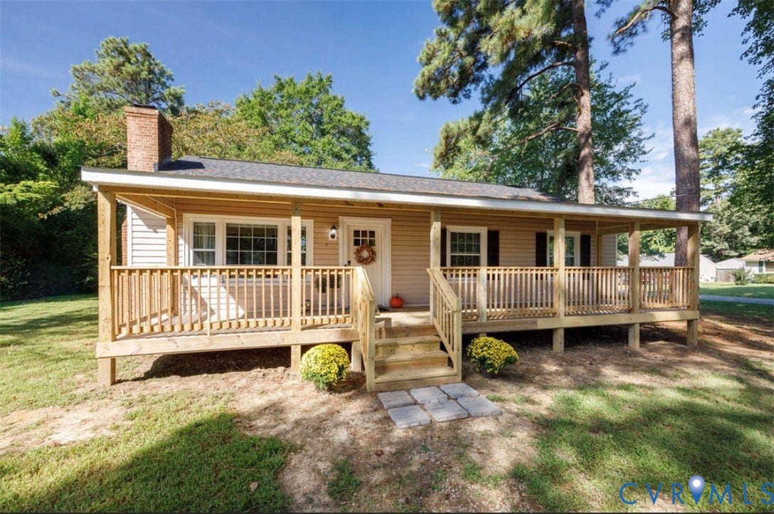 Expansive front porch with natural wood railing, light-toned siding, dark shutters, and a brick chimney