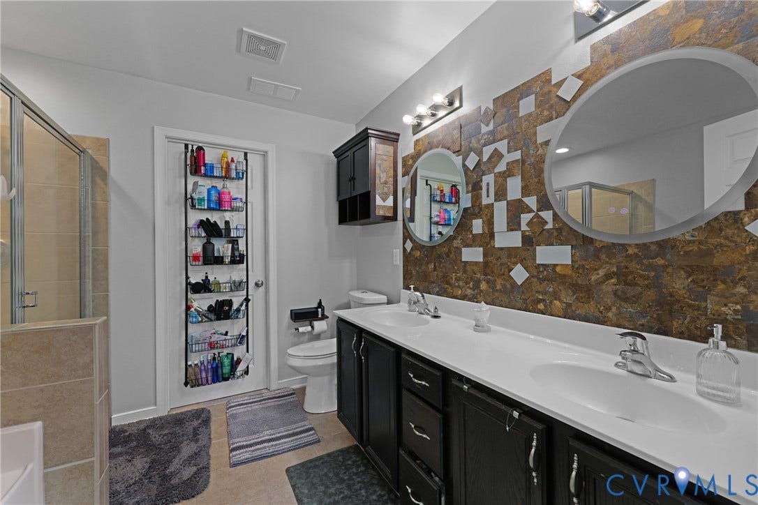Dual vanity with individual sinks, decorative tile wall accent, round and oval mirrors, dark wood-finish cabinetry, and a walk-in shower with glass enclosure