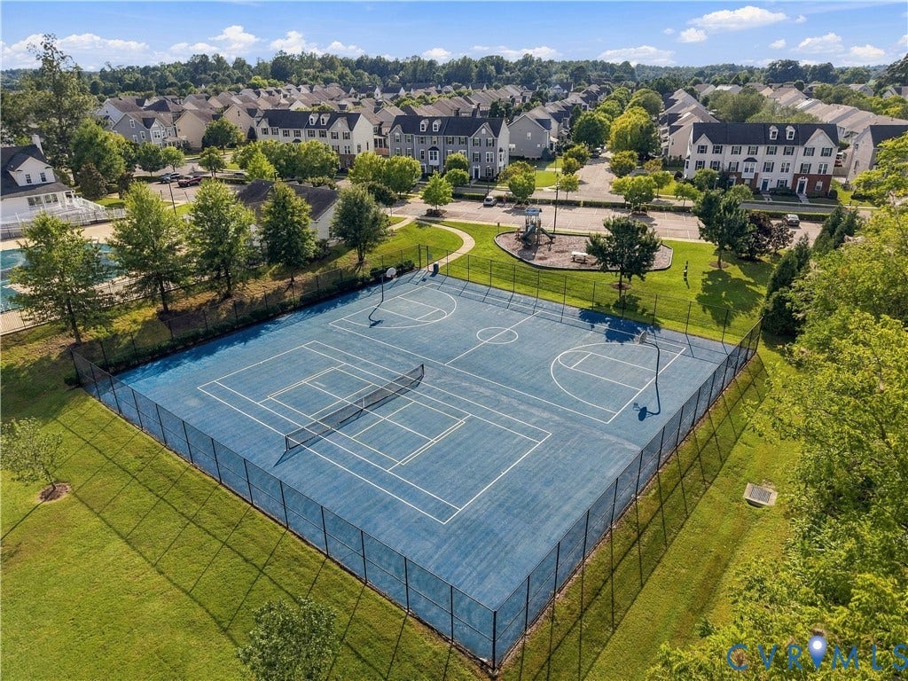 Fenced multi-sport court with basketball hoops and tennis net, bordered by green lawn and mature trees