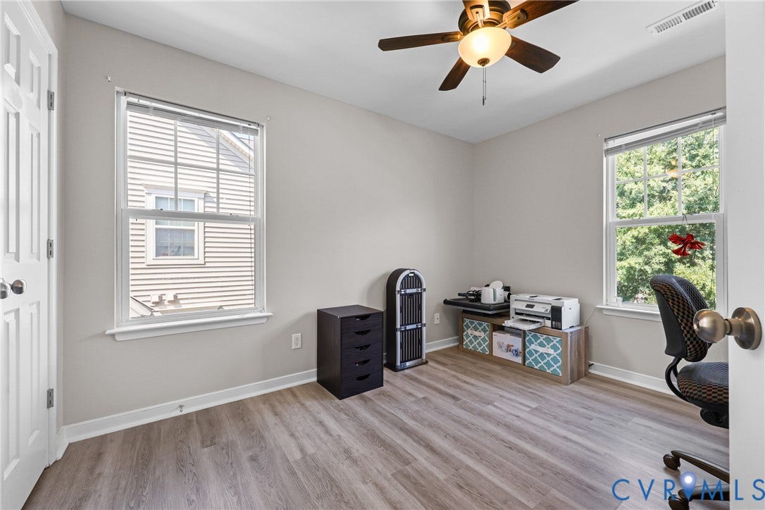 Wood-finish flooring, two large windows with blinds, ceiling fan with integrated lighting, neutral wall paint, and white trim throughout
