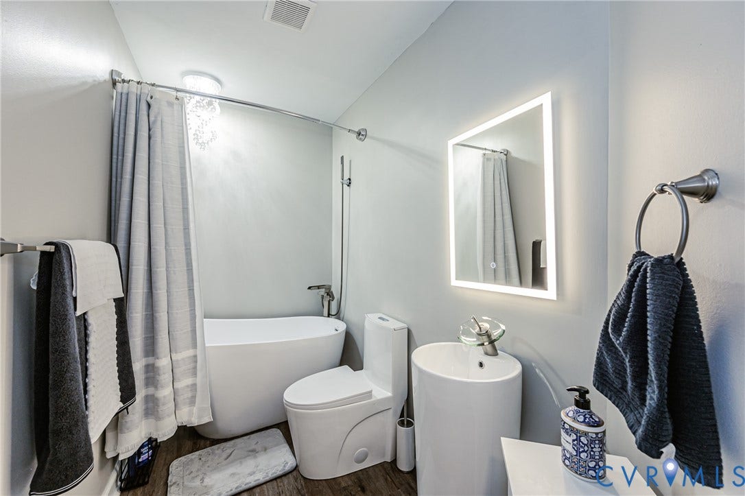 Freestanding soaking tub, wood-finish flooring, illuminated vanity mirror, pedestal sink, and modern toilet