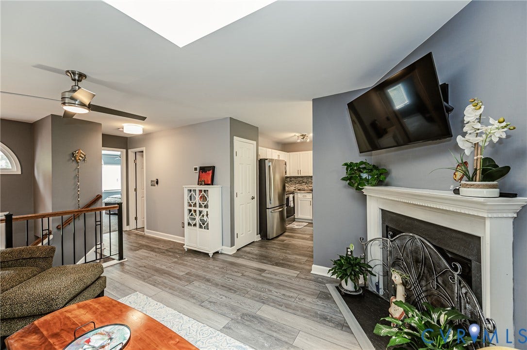 Wood-finish flooring, fireplace with white mantel, ceiling fan with integrated lighting, skylight, and stainless steel appliances in the kitchen
