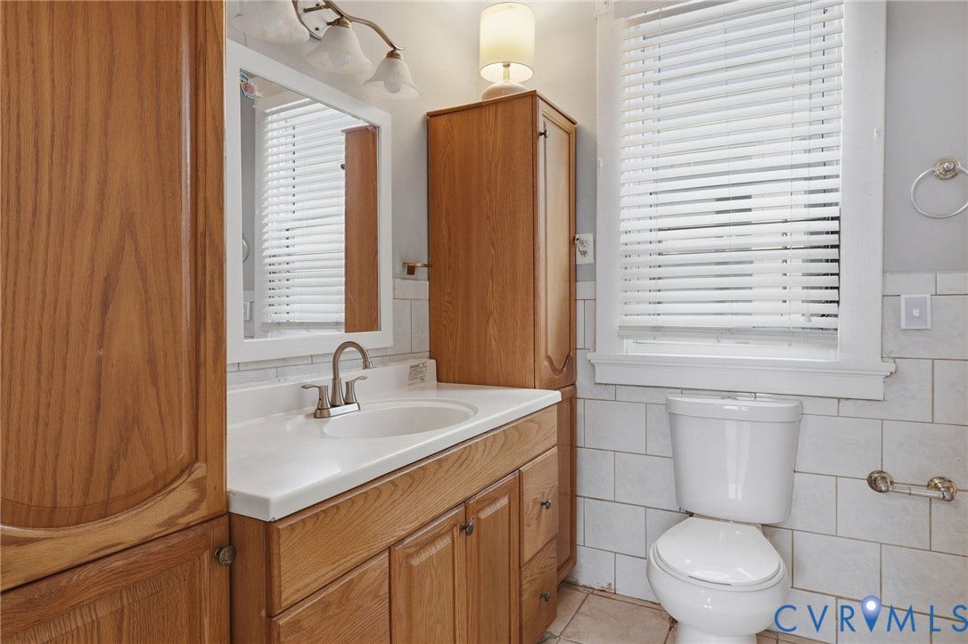 Wood-finish cabinetry, white tile walls, white vanity countertop with integrated sink, and brushed nickel fixtures
