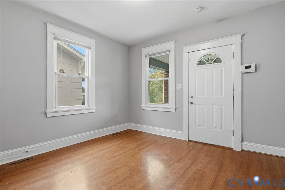 Wood-finish flooring, light gray wall paint, two pane windows with white trim, and a paneled entry door with an arched transom