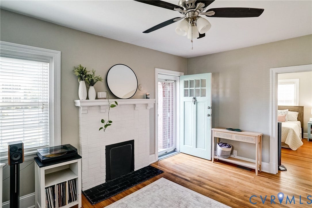 Hardwood flooring, painted brick fireplace with hearth, ceiling fan, natural light from window, and light-colored entry door with glass panels
