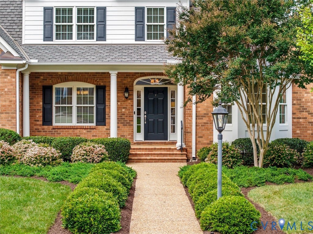 Brick exterior with white siding accents, covered entry with columns, dark entry door, and manicured landscaping with a paved walkway