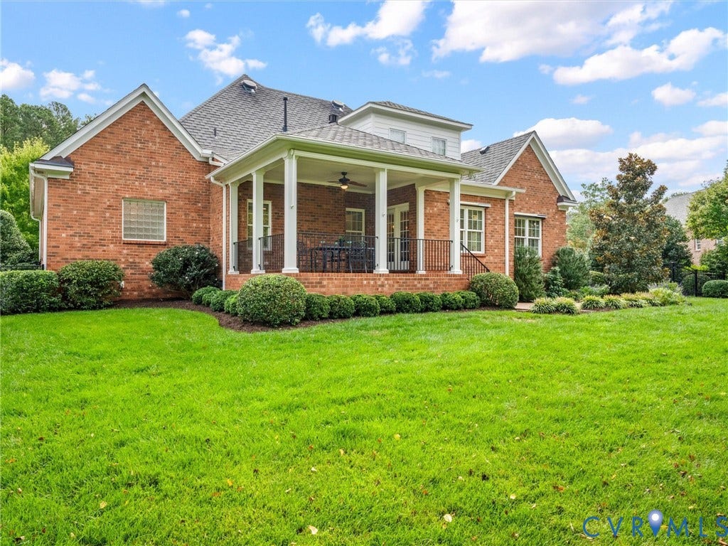 Expansive green lawn, brick exterior, covered patio with white columns, black railing, and mature landscaping