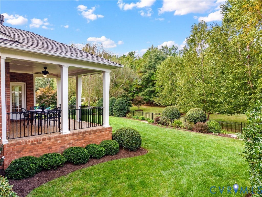 Brick exterior with a covered patio, white columns, ceiling fan, and fenced yard with mature landscaping