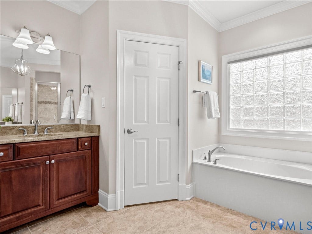 Built-in bathtub with deck-mounted faucet, glass block window, wood vanity with undermount sink, and tiled flooring