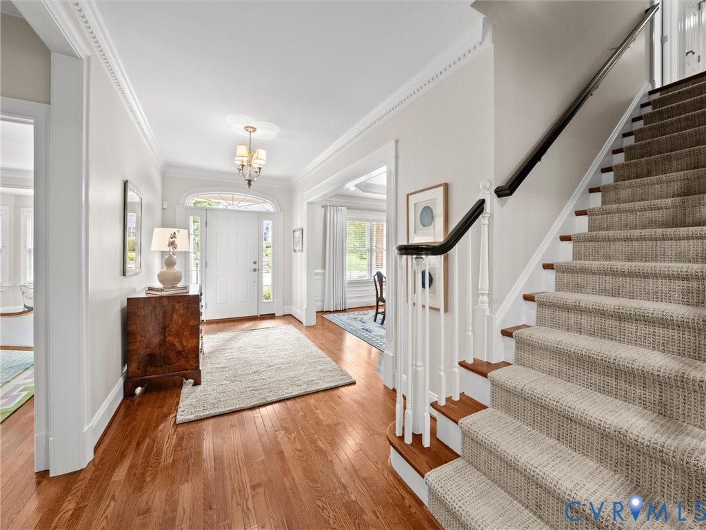 Hardwood flooring, white paneled entry door with arched transom, staircase with carpeted treads and dark wood handrails, decorative crown molding, and an overhead chandelier