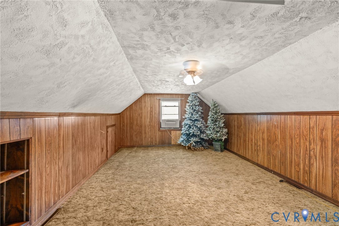 Sloped ceiling with textured finish, wood-paneling walls, built-in shelving, window unit, and ceiling fan with light kit